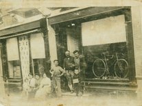 Magasin de cycles Pautric, sur le pas de la porte André Pautric avec peut-être son père à côté de lui, la dame qui tient la petite-fille sur les genoux est sa soeur Marie Photo de magasin de cycles